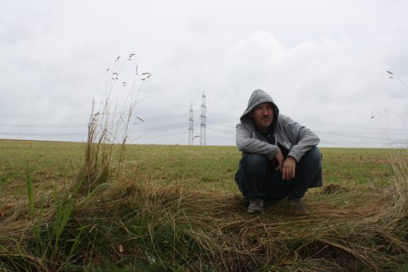 Mann kniet in einem Feld mit grauem Himmel und einem Strommast im Hintergrund.