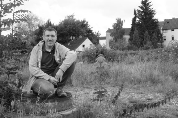 Mann sitzt auf einer Steinplatte in einer bewachsenen Umgebung mit Häusern im Hintergrund.