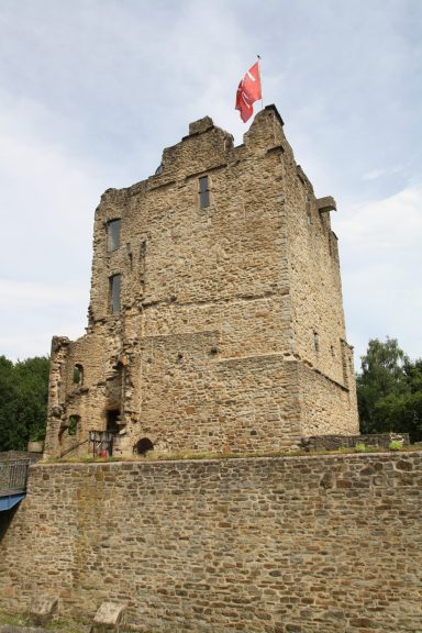 Mittelalterliche Steinhäuser mit einer roten Fahne, umgeben von Bäumen und blauem Himmel.