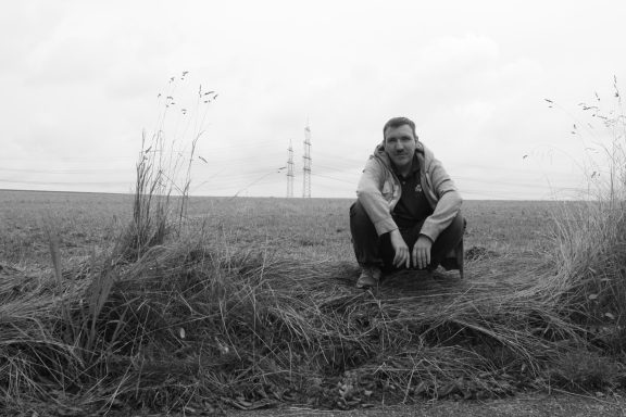 Mann sitzt auf dem Boden in einer düsteren, offenen Landschaft mit Strommasten im Hintergrund.