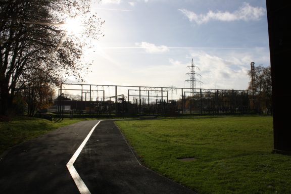 Blick auf ein freies Feld mit einer Stromleitung und einem Industriegebäude im Hintergrund.