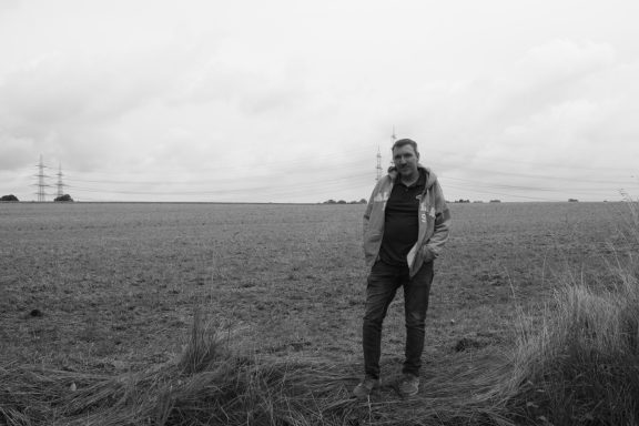 Mann steht in einem weiten, leeren Feld unter einem bewölkten Himmel.