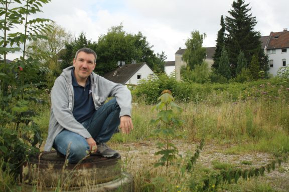 Mann kniet in einer offenen, bewachsenen Fläche mit Gebäuden im Hintergrund.