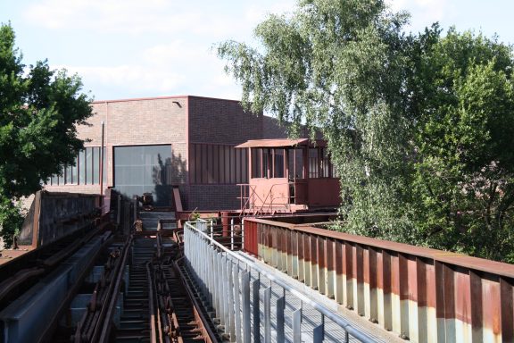 Verlassene Gleise mit einem alten Waggon und einem angrenzenden Gebäude, umgeben von Bäumen.