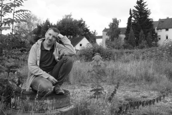 Mann sitzt nachdenklich in einer verwilderten Gartenlandschaft.